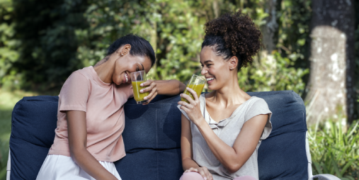 Fotografía de dos mujeres sentadas en un sofá, bebiendo jugo y riendo y, al fondo, varios árboles