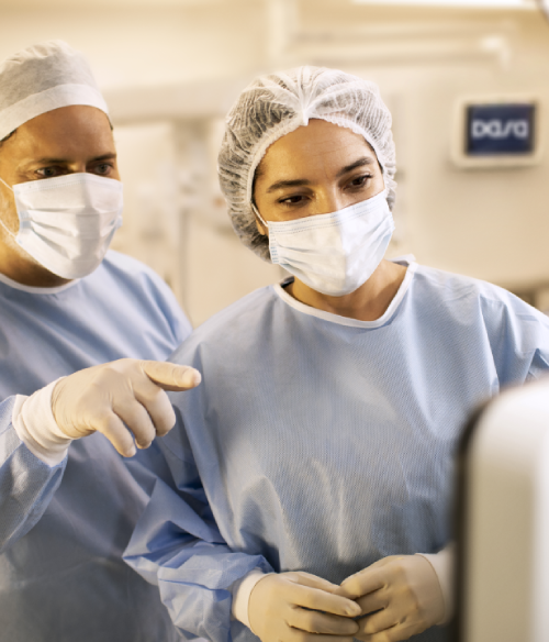 Um homem e uma mulher, usando toucas, máscaras, luvas e aventais, olhando para um
monitor em uma sala com outros equipamentos médicos